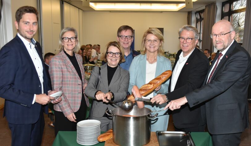 Handwerkspräsident, Kreishandwerkermeister, Geschäftsführer und drei Damen als Gäste stehen um einen Topf mit Würstchen und Brot